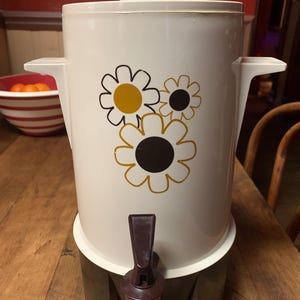 May include: White vintage coffee pot with a brown base and a spigot. The pot has a floral design with three yellow and black daisies.
