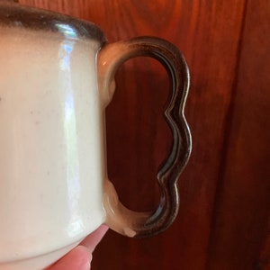 May include: A white ceramic pitcher with a brown handle. The handle has a scalloped edge.