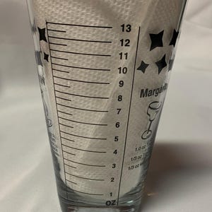 Puede incluir: Un vaso medidor de vidrio transparente con marcas negras para onzas. El vaso tiene un dise&ntilde;o de estrella negra y el texto "Margarita" impreso en el lateral. El vaso est&aacute; marcado con medidas de 1 a 13 onzas.