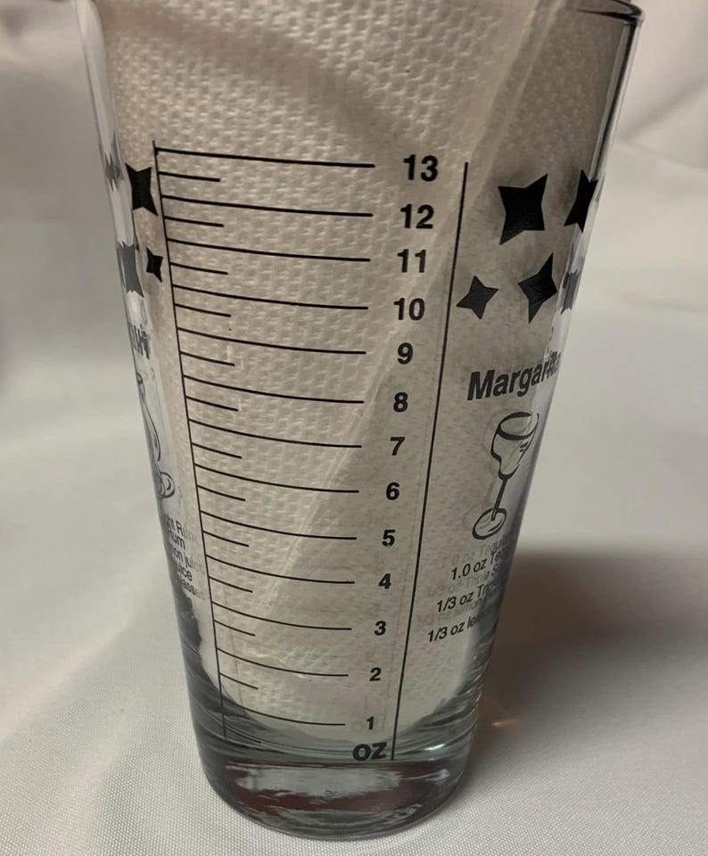 Puede incluir: Un vaso medidor de vidrio transparente con marcas negras para onzas. El vaso tiene un dise&ntilde;o de estrella negra y el texto "Margarita" impreso en el lateral. El vaso est&aacute; marcado con medidas de 1 a 13 onzas.