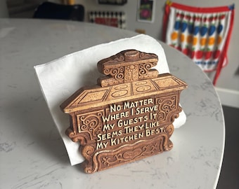 May include: A decorative wooden napkin holder with ornate carvings and the text "NO MATTER WHERE I SERVE MY GUESTS IT SEEMS THEY LIKE MY KITCHEN BEST." The holder is brown and sits on a white napkin.