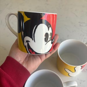 K&ouml;nnte beinhalten: Drei Keramikbecher mit Mickey Mouse. Ein Becher, der in einer Hand gehalten wird, zeigt Mickys Gesicht vor einem roten, schwarzen und gelben Hintergrund. Die anderen beiden Becher sind sichtbar, einer mit Mickys Hand und der andere ist schlicht wei&szlig;.