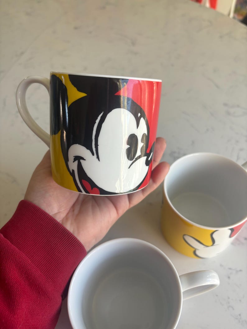 K&ouml;nnte beinhalten: Drei Keramikbecher mit Mickey Mouse. Ein Becher, der in einer Hand gehalten wird, zeigt Mickys Gesicht vor einem roten, schwarzen und gelben Hintergrund. Die anderen beiden Becher sind sichtbar, einer mit Mickys Hand und der andere ist schlicht wei&szlig;.