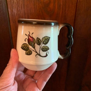 May include: A white ceramic mug with a brown rim and handle. The mug has a floral design with a single rose and leaves in shades of green and red.