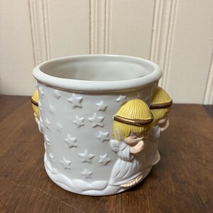 May include: White ceramic planter with embossed stars and cherub figures. The planter has a round shape with a wide opening and a decorative base. The cherubs have yellow hair and gold halos. Suitable for small plants.