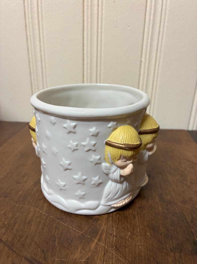 May include: White ceramic planter with embossed stars and cherub figures. The planter has a round shape with a wide opening and a decorative base. The cherubs have yellow hair and gold halos. Suitable for small plants.