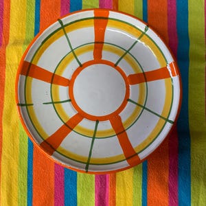 May include: A white ceramic plate with a circular design of orange, yellow, and green stripes. The plate is on a colorful striped tablecloth.