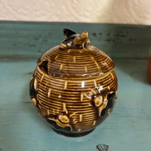 May include: A brown ceramic honey pot shaped like a beehive with a bee on top. The pot is decorated with bees and has a brown and white striped pattern.