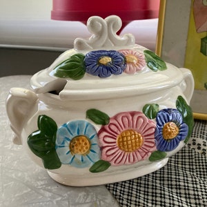 May include: White ceramic serving dish with a lid. The lid has a floral design with blue, pink, yellow, and green flowers. The dish has a similar floral design with blue, pink, and yellow flowers.