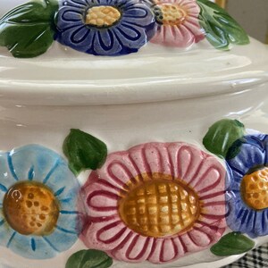 May include: White ceramic cookie jar with a floral design. The jar has a lid with a handle and is decorated with pink, blue, and yellow flowers.