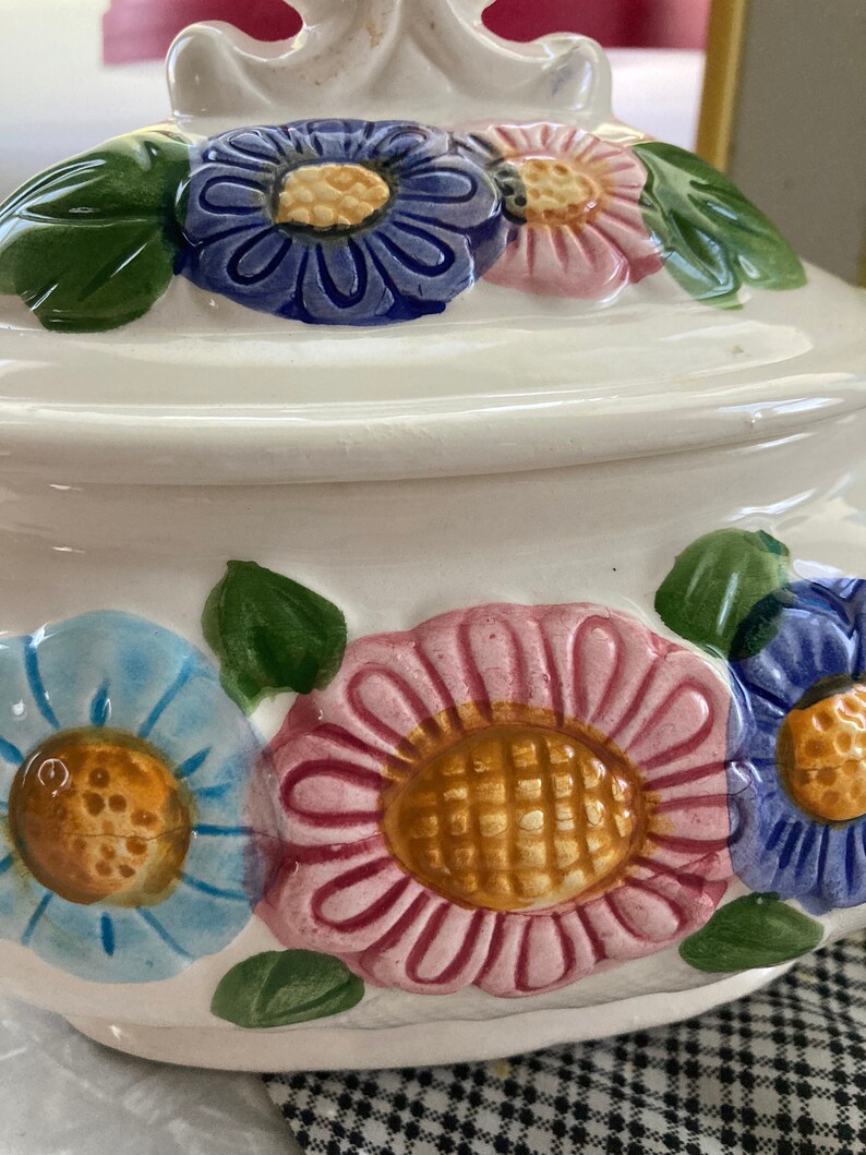 May include: White ceramic cookie jar with a floral design. The jar has a lid with a handle and is decorated with pink, blue, and yellow flowers.
