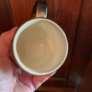 May include: A brown and white ceramic mug with a rounded handle. The mug is empty and has a white interior.