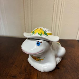 May include: A white ceramic frog figurine featuring a removable hat lid decorated with a yellow sunflower. The frog has blue eyes, long eyelashes, a pink mouth, and a yellow sunflower painted on its chest. The surface is glossy and smooth.
