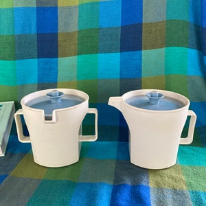 May include: Two white ceramic creamer and sugar containers with blue lids and handles. The containers are on a blue and green checkered tablecloth.