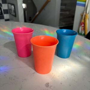 May include: Three colorful plastic cups in pink, orange, and blue are arranged on a white countertop. The cups are empty and have a simple, straight-sided design. The lighting creates a rainbow effect on the surface.