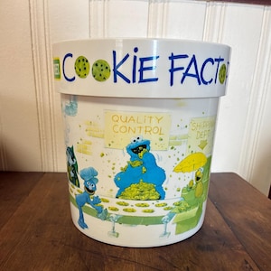 May include: White plastic cookie container with the words "Cookie Factory" in blue and green. The container features cartoon characters and the words "Quality Control" and "Shipping Dept."