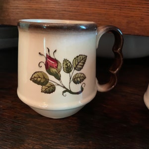 May include: A white ceramic mug with a brown rim and handle. The mug features a hand-painted design of a single rose bud with green leaves.