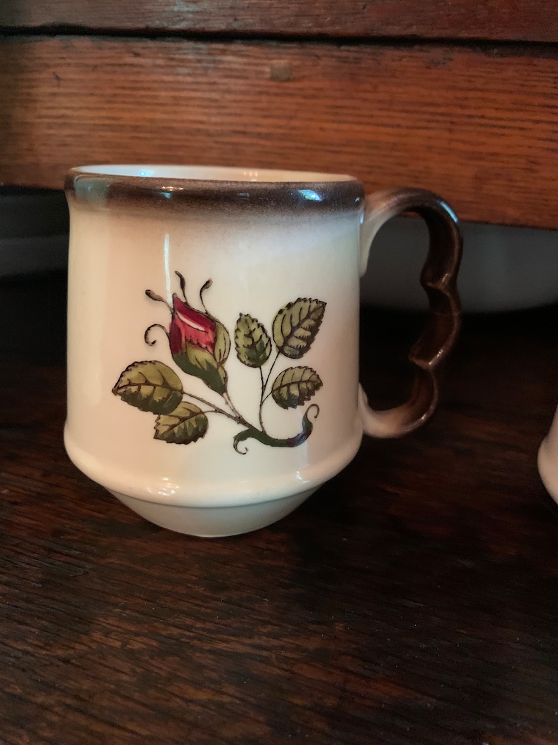 May include: A white ceramic mug with a brown rim and handle. The mug features a hand-painted design of a single rose bud with green leaves.