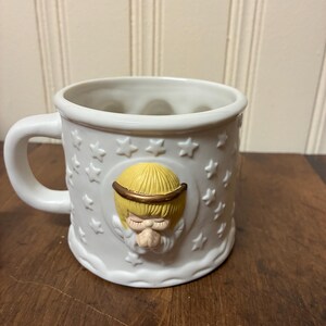 May include: White ceramic mug with a handle, decorated with embossed stars and an angel with a yellow halo and hair. The angel's face is in relief, giving a three-dimensional effect to the mug's design.