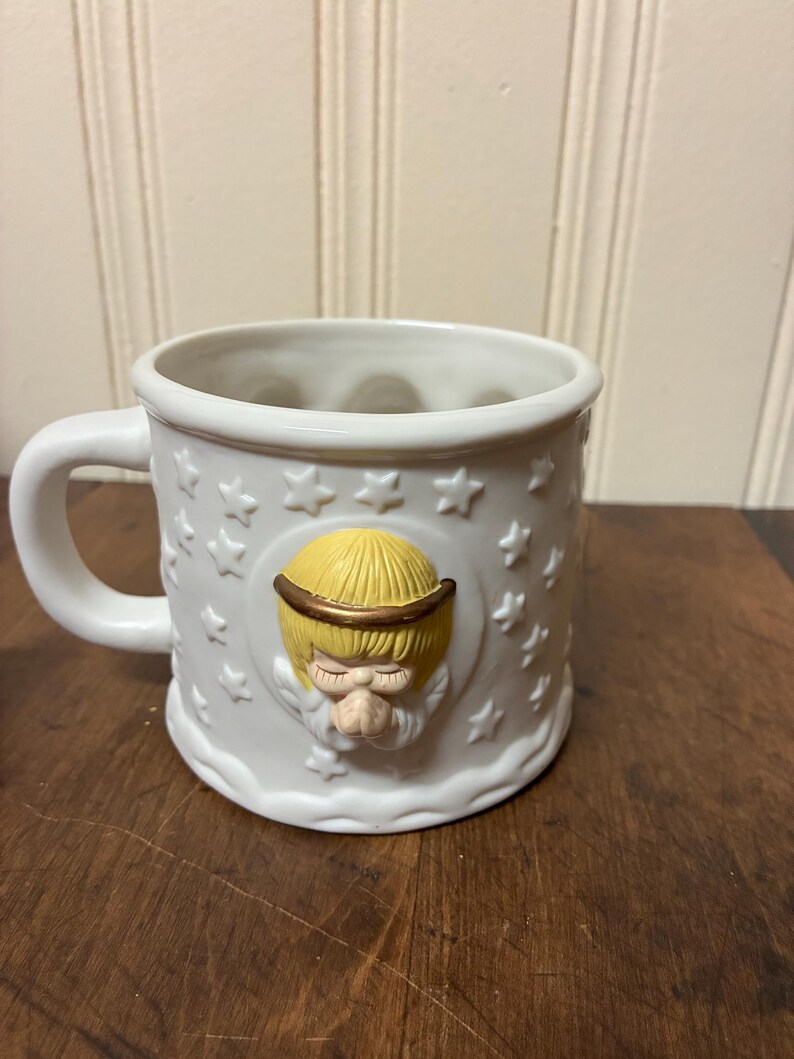 May include: White ceramic mug with a handle, decorated with embossed stars and an angel with a yellow halo and hair. The angel's face is in relief, giving a three-dimensional effect to the mug's design.