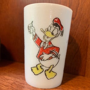 May include: White ceramic mug with a cartoon image of Donald Duck wearing a red and white shirt and a red hat. The mug has a textured surface.