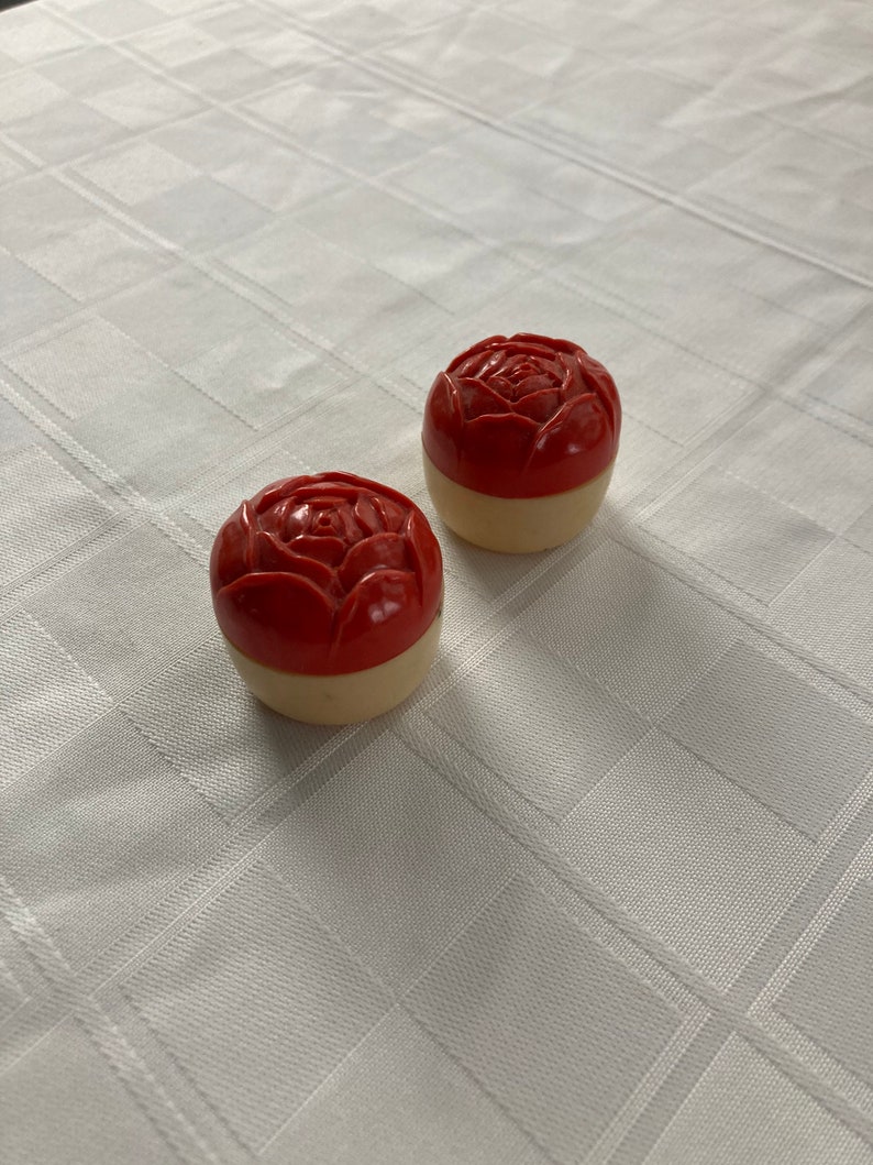 May include: Two small, round, red and white containers with a rose design on the top. The containers are made of plastic and have a vintage look.