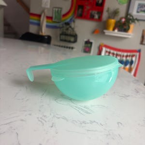 May include: A light teal plastic measuring bowl with a handle and a matching lid. The bowl has a rounded shape and is sitting on a white surface. The handle is curved and extends from the side of the bowl.
