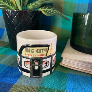 May include: White ceramic mug with a black and red design of a diner with the text "Big City Diner Open 24 Hours".
