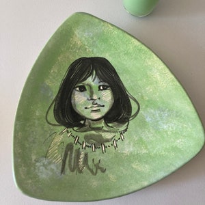 May include: A green ceramic plate with a painted image of a woman's face. The plate is triangular in shape and has a textured surface.