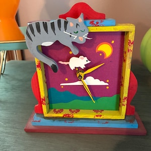 May include: A whimsical wooden clock featuring a gray cat and white mouse design. The clock face has a colorful landscape with a yellow moon and clouds. The clock is painted with red, yellow, blue, and purple accents.