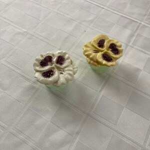May include: Two ceramic flower-shaped bottle stoppers. One is white with purple centers and the other is yellow with brown centers.