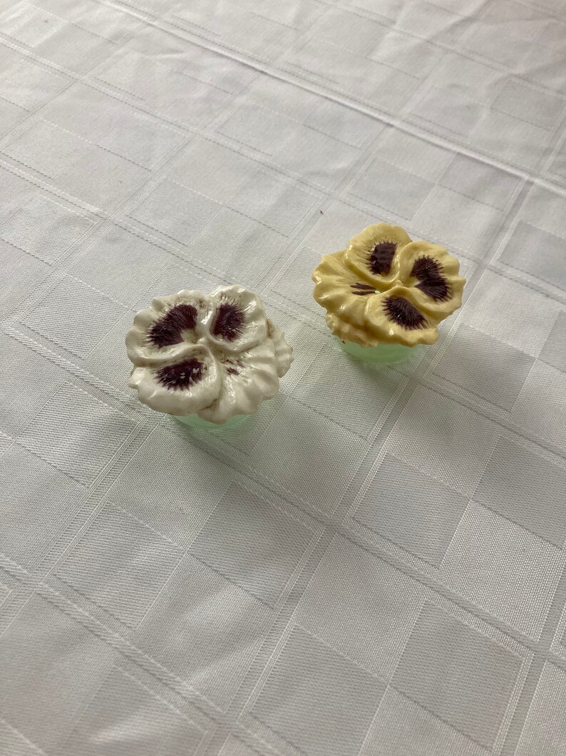 May include: Two ceramic flower-shaped bottle stoppers. One is white with purple centers and the other is yellow with brown centers.