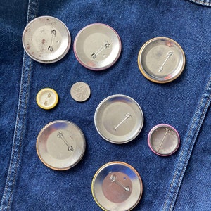 May include: Nine different button pins with various colors and designs. The pins are on a blue denim background.