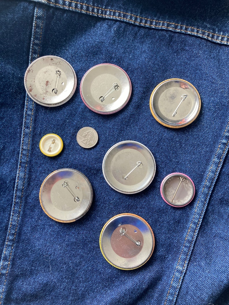 May include: Nine different button pins with various colors and designs. The pins are on a blue denim background.