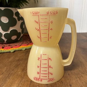 May include: A vintage, two-part, pale yellow measuring cup with a handle. Red markings indicate measurements in ounces and cups. The cup is made of a translucent plastic material, and the measurements are clearly visible.