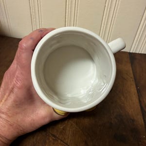 May include: A white ceramic mug with a small handle. The mug is held in a person's hand, and the interior is visible. The mug sits on a wooden surface.