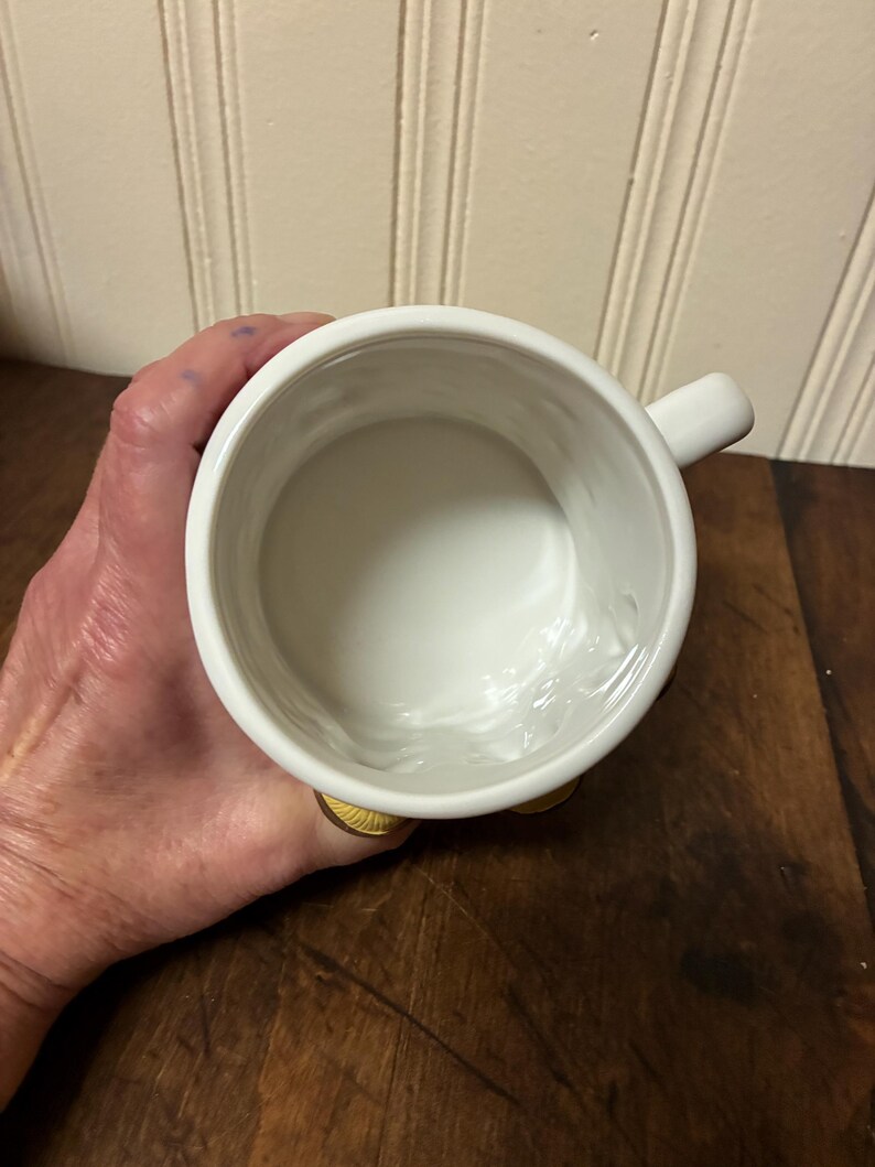 May include: A white ceramic mug with a small handle. The mug is held in a person's hand, and the interior is visible. The mug sits on a wooden surface.