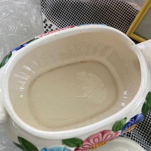 May include: A white ceramic serving dish with a floral design. The dish has a large, oval-shaped bowl with a smaller, round bowl on top. The floral design features pink, blue, and yellow flowers with green leaves.