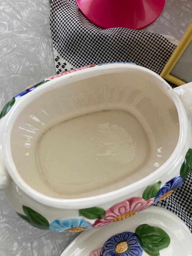 May include: A white ceramic serving dish with a floral design. The dish has a large, oval-shaped bowl with a smaller, round bowl on top. The floral design features pink, blue, and yellow flowers with green leaves.