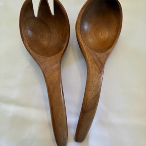 May include: A set of two wooden salad servers. The server on the left has a fork-like head and the server on the right has a spoon-like head. Both servers are made of light brown wood.