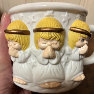 May include: White ceramic mug with three-dimensional angel figures. The angels have yellow hair and gold halos, and are in a praying pose. The mug has a handle and is decorated with star shapes. The mug is suitable for hot beverages.