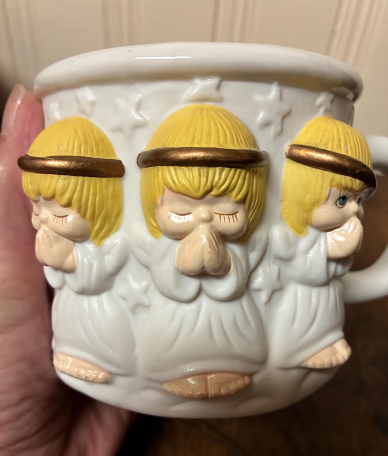 May include: White ceramic mug with three-dimensional angel figures. The angels have yellow hair and gold halos, and are in a praying pose. The mug has a handle and is decorated with star shapes. The mug is suitable for hot beverages.
