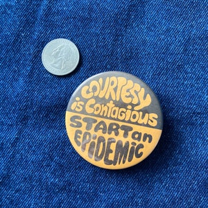 May include: A round button with a black and gold design. The text on the button reads "Courtesy is contagious. Start an epidemic."