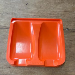 May include: An orange Tupperware spoon rest with two recessed areas. The rectangular rest has rounded edges and the Tupperware logo is visible at the bottom. The plastic is a bright, solid orange color.