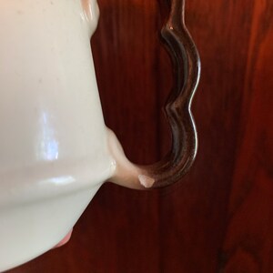 May include: Close-up of a white ceramic teapot with a brown handle. The handle has a scalloped edge and is slightly chipped.