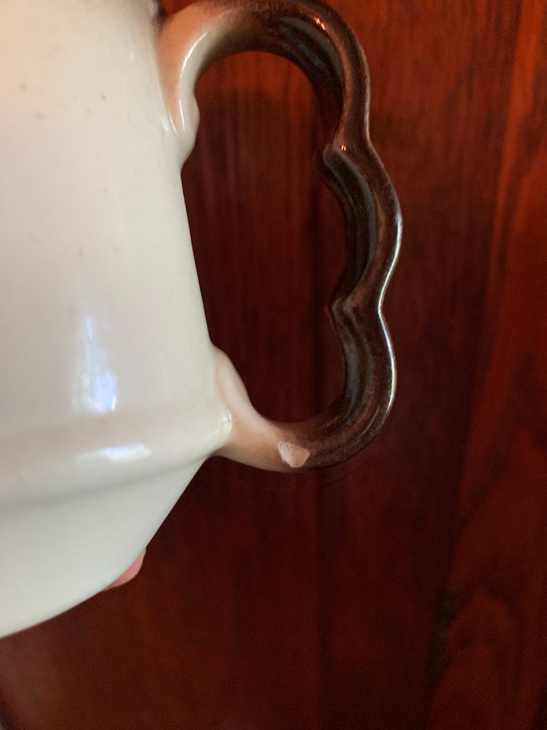 May include: Close-up of a white ceramic teapot with a brown handle. The handle has a scalloped edge and is slightly chipped.
