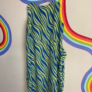May include: A sleeveless maxi dress with a blue and green wavy pattern. The dress is hanging on a hanger against a wall with a rainbow painted on it.