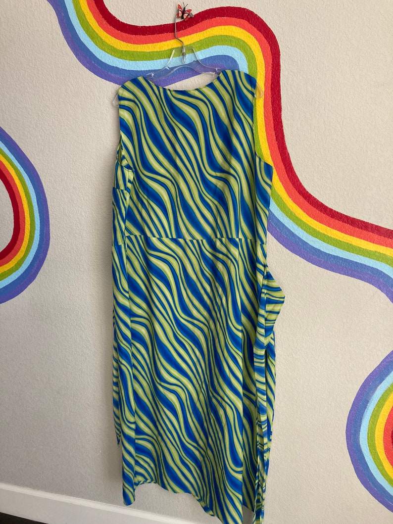 May include: A sleeveless maxi dress with a blue and green wavy pattern. The dress is hanging on a hanger against a wall with a rainbow painted on it.