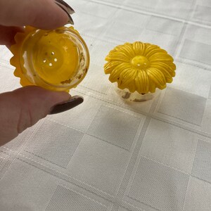 May include: Two yellow plastic salt and pepper shakers shaped like sunflowers. The shakers have a clear plastic base and a yellow plastic sunflower top.