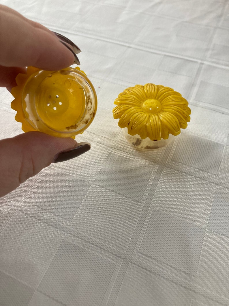 May include: Two yellow plastic salt and pepper shakers shaped like sunflowers. The shakers have a clear plastic base and a yellow plastic sunflower top.